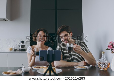 Young happy couple two woman man in casual t-shirt clothes sit by table eat pancakes talk video call mobile cell phone drinking coffee cook food in light kitchen at home together Healthy diet concept.