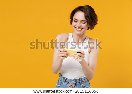 Young gambling woman 20s with bob haircut wear white tank top shirt using play racing on mobile cell phone hold gadget smartphone for pc video games isolated on yellow color background studio portrait