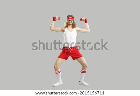 Full body skinny nerd with funny face doing exercise with dumbbells isolated on gray background. Full length hilarious smiling man in sweatband, white tank top and red shorts enjoying sports workout