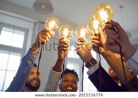 Happy smiling interracial group of people standing together with raised lightbulb office shot. Focus on illuminated glowing lamp. Creative thinking and teamwork, brainstorming and idea share concept Royalty-Free Stock Photo #2015156774