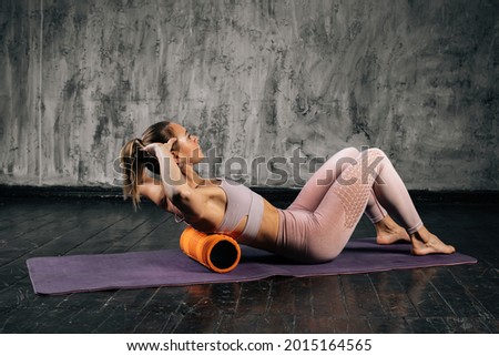 Muscular young athletic woman with perfect beautiful body in activewear using foam roller massager on upper back lying on yoga mat. Caucasian fitness female posing in studio with dark grey background.
