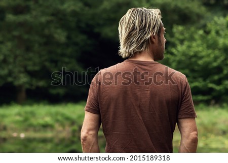 Blonde man in brown t-shirt stands in a forest near a pond. Rear view.