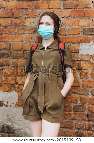 schoolgirl poses against a brick wall in the backyard of the school, wearing a protective mask on her face from a coronavirus infection, education and back to school concept