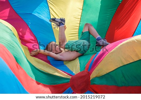 Happy boy having fun and waving in the center of bright colorful parachute, outdoors party, summer school camp, healthy active leisure, childhood and emotions