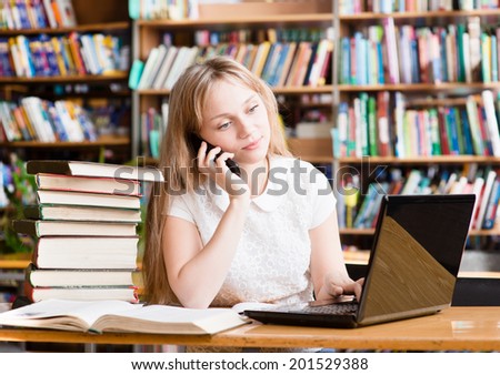 Young female student in library typing on laptop and talking on the phone