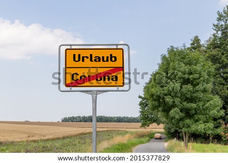 Yellow street sign or town sign with Corona and vacation written in German. and Corona is crossed out and in the background you see a car driving. 