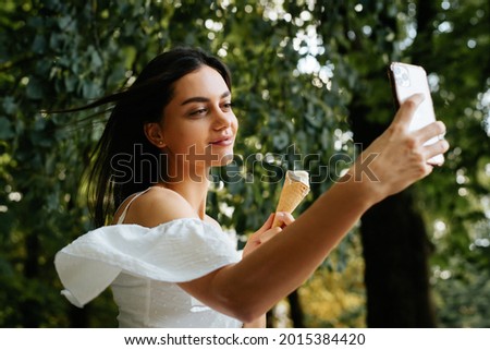 Happy young indian brunette woman in white dress taking selfie on smartphone, holding ice cream and resting at weekend in park on summer day. Smiling beautiful woman enjoying vacation, lifestyle