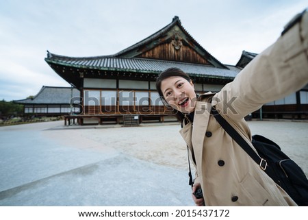 joyful korean visitor is taking a selfie with traditional religious building in Kyoto. excited female tourist looking at camera is taking picture to commemorate her trip to Nijo Castle.