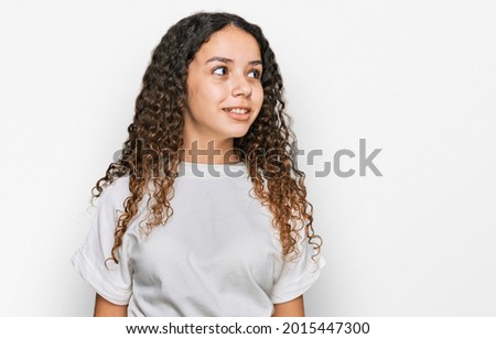 Teenager hispanic girl wearing casual white t shirt looking away to side with smile on face, natural expression. laughing confident. 