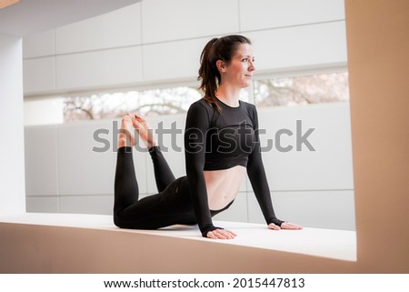 Young flexible yoga woman in black practice cobra asana on yoga mat, wearing black sportswear, in studio indoor over panoramic window on reflective modern background 