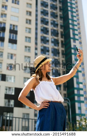 Young beautiful woman blogger in summer clothing making selfie in front of modern building. Vertical format, modern culture, sunny day, city view. Woman is wearing hat