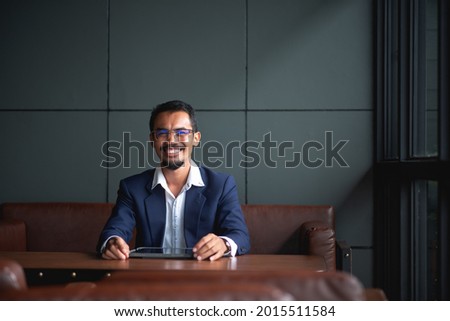 Young handsome businessman wears glasses and works on touchpad while sitting on sofa in office.