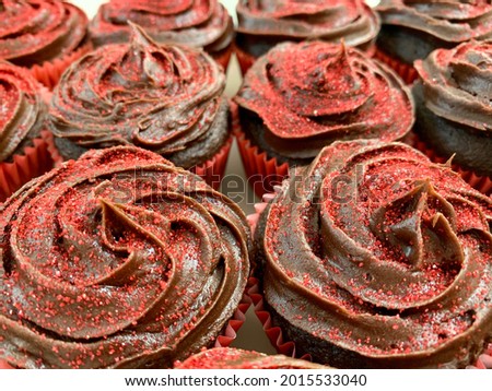 The top down, close up view of a tray of cupcakes. The sweet treats fully fill the frame in this delicious background. They are chocolate cupcakes, with fudge frosting, and bright red sprinkles.