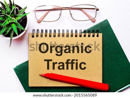On a light background, gold-framed glasses, a flower in a pot, a green notebook, a red pen and a brown notebook with the text ORGANIC TRAFFIC. Business concept
