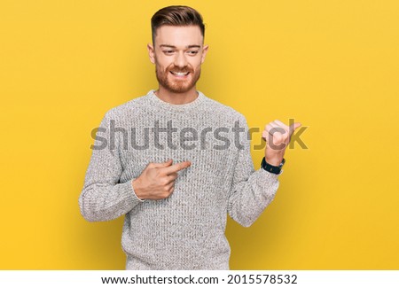 Young redhead man wearing casual winter sweater pointing to the back behind with hand and thumbs up, smiling confident 