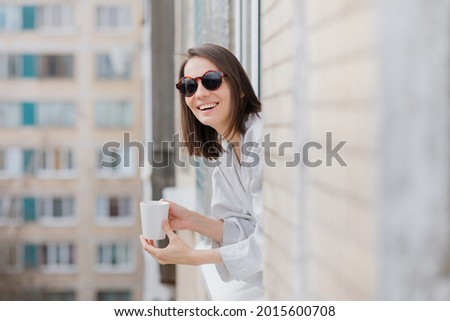 European woman in sunglasses and with a mug of coffee or tea looking out of the window and smiling, quarantine and self-isolation in a covid-19, young and beautiful woman looking out of the window and