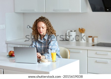 Enjoying time at home. Beautiful young smiling woman working on laptop and drinking orange juice while sitting on the chair in the kitchen at home. Young woman working at home in the morning Royalty-Free Stock Photo #2015619038