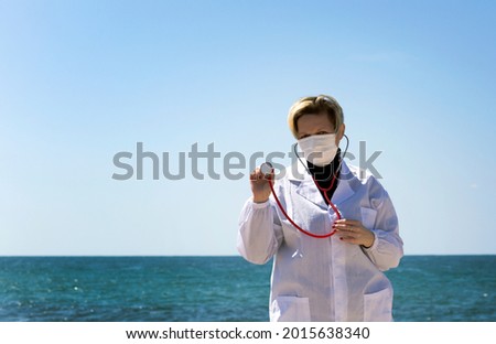 Blond doctor woman of Russian Caucasian appearance Holding A Stethoscope In Hand. Portrait of professional confident American doctor in medical mask and white coat, stethoscope over neck on a beach