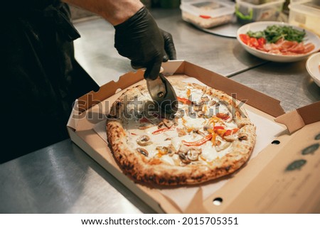 Close-up image. Chef in restaurant cutting round tasty pizza with vegetables into pieces in cardboard box. Fresh hot favourite dish. Concept of cooking, food preparation, restaurant work.