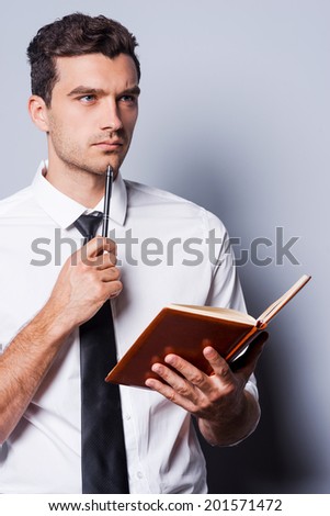 Waiting for inspiration. Thoughtful young man in shirt and tie holding note pad and touching his chin with pen while standing against grey background