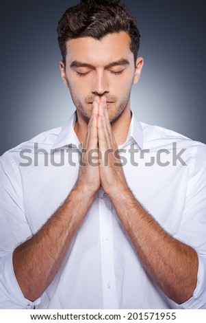 Praying for business. Handsome young man in white shirt holding hands clasped near face while standing against grey background
