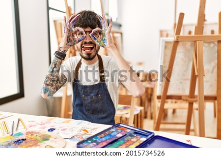 Hispanic man with beard at art studio doing ok gesture like binoculars sticking tongue out, eyes looking through fingers. crazy expression. 