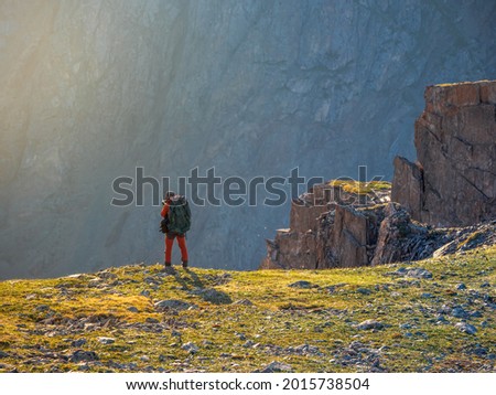 Photographer with a large backpack takes pictures of a beautiful mountain landscape on the edge of a cliff. Dangerous mountains and abyss in sunset. 