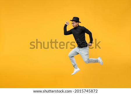 Full length side view of young active sporty fashionable fun african man 20s wear stylish black hat shirt eyeglasses jump high run fast hurrying isolated on yellow orange background studio portrait.