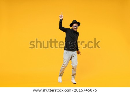 Full length young fun african man 20s wearing stylish black hat shirt eyeglasses doing winner gesture pointing index finger up walk going isolated on yellow orange color background studio portrait. Royalty-Free Stock Photo #2015745875