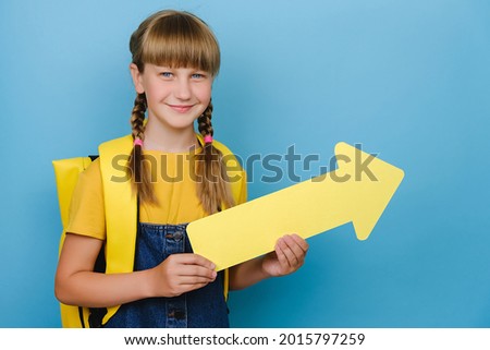 Portrait of smiling blonde schoolgirl showing aside with yellow arrow, wears backpack, posing isolated over blue color background wall with copy space for promotional content. Back to school concept
