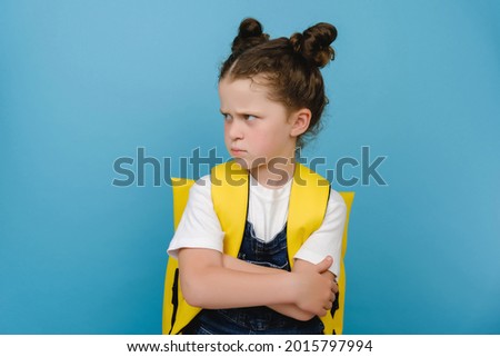 Portrait of angry sad little schoolgirl, crossed arms looking to side, does not want to go to school, wears yellow backpack, posing isolated over blue studio background. Children's emotions concept Royalty-Free Stock Photo #2015797994