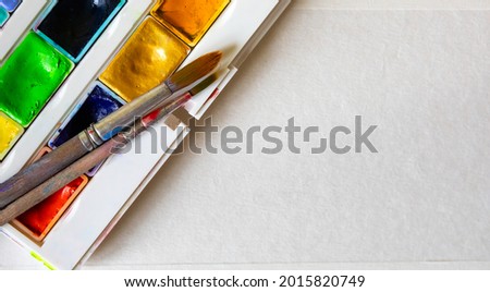 Watercolors in cuvettes in a box and brushes for painting on a white sheet of paper. Blank for text, banner, design