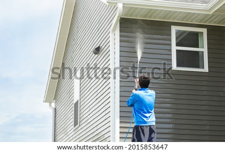 Man in blue jacket cleans dusk and dirt from exterior siding and under roof with a high-pressure nozzle spray with water soap cleaner. Wash a house during the day. Home maintenance service concept. Royalty-Free Stock Photo #2015853647