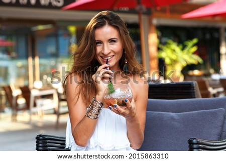 Elegant fashionable brunette woman drinking alcoholic tasty cocktail and having fun, wearing cute white beach dress and trendy accessorizes . Royalty-Free Stock Photo #2015863310