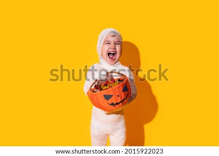 Child in mummy costume holding basket of chocolates in front of yellow background. Royalty-Free Stock Photo #2015922023