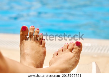 Beautiful legs of a girl with a pink manicure. Young woman is lying near the pool with blue water on a chaise longue