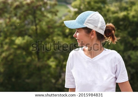 Young woman in a white T-shirt and baseball cap on her head looks away on a background of greenery