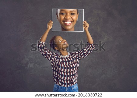 Happy pretty young black woman with short hair holding photo of her face with clear skin and perfect charming smile standing against dark grey studio background. Human emotions and dental care concept