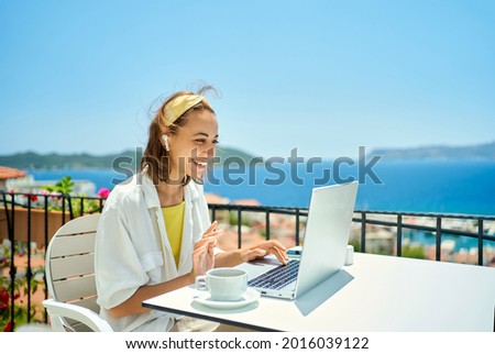 beautiful caucasian woman in wireless earphones talk speak on video call on computer while sitting at balcony with seaview on resort. female have online conversation, work chat conference or Royalty-Free Stock Photo #2016039122