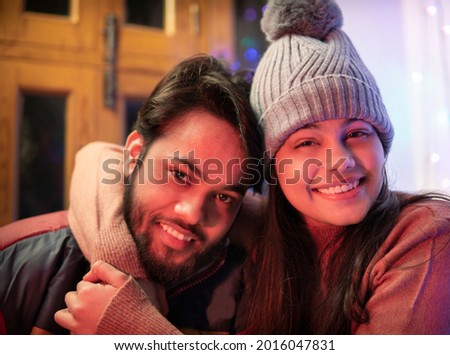 Happy Indian brother and sister sitting together at home in winters and embracing each other with a toothy smile and looking at the camera. They are wearing warm clothes, sweaters, jacket andcap. Royalty-Free Stock Photo #2016047831