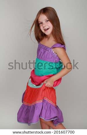 Studio image of a charming little girl with long healthy hair and bright summer sundress dancing and smiling at the camera on a gray background