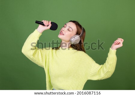 Young woman with perfect natural makeup brown big lips in casual sweater on green background with wireless headphones hold  microphone singing