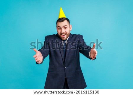 Come into my arms! Friendly man in official style suit and party cone giving free hugs with outstretched hands, welcoming inviting to embrace. Indoor studio shot isolated on blue background.