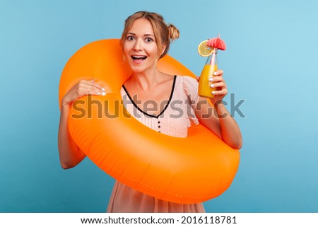 Excited woman with funny hair buns relaxing at summer resort, holding orange rubber ring and enjoying fresh fruit cocktail, laughing happily at vacation. Indoor studio shot isolated on blue background