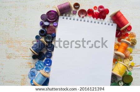 Rainbow spectrum buttons and reels with colorful threads, laid  around a notebook with space for text on a light wooden background. Hobbies and fashion lessons concepts