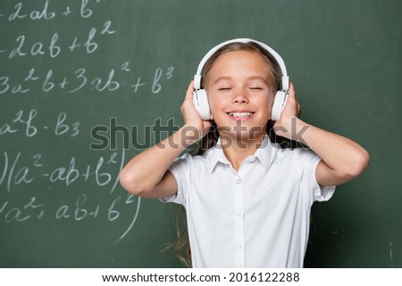 joyful schoolgirl with closed eyes listening music in headphones near chalkboard