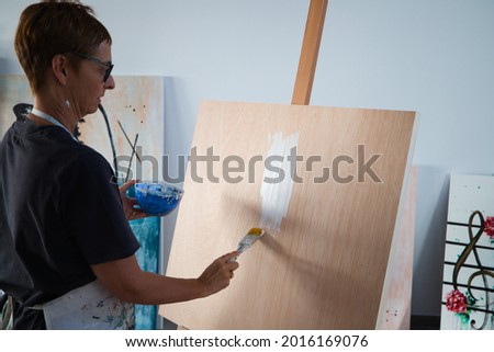 Mature woman with glasses is painting a white background to make a new work of art in her studio. The canvas is on the easel. Concept art and artist.