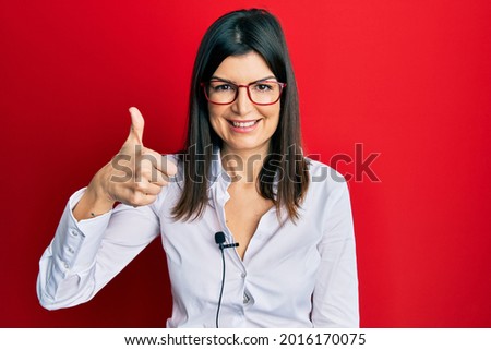 Young hispanic woman using lavalier microphone smiling happy and positive, thumb up doing excellent and approval sign 