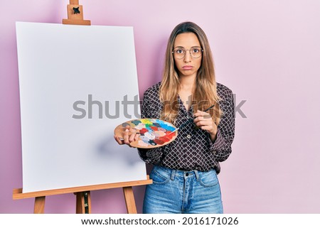Beautiful hispanic woman standing drawing with palette by painter easel stand depressed and worry for distress, crying angry and afraid. sad expression. 
