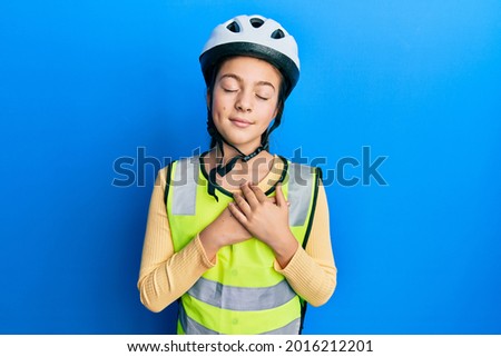 Beautiful brunette little girl wearing bike helmet and reflective vest smiling with hands on chest with closed eyes and grateful gesture on face. health concept. 
